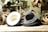 A collection of hats is displayed on a wooden surface, surrounded by loops of material. The hats are primarily dark in color, with one lighter hat showing its underside. The setting appears to be a workshop, as suggested by the blurred background with a person working and various tools visible.
