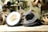 A collection of hats is displayed on a wooden surface, surrounded by loops of material. The hats are primarily dark in color, with one lighter hat showing its underside. The setting appears to be a workshop, as suggested by the blurred background with a person working and various tools visible.