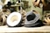 A collection of hats is displayed on a wooden surface, surrounded by loops of material. The hats are primarily dark in color, with one lighter hat showing its underside. The setting appears to be a workshop, as suggested by the blurred background with a person working and various tools visible.