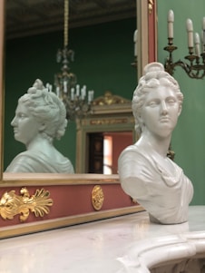 A marble bust of a woman is placed on a polished surface in front of a decorative mirror with intricate gold accents. In the background, an ornate chandelier and green wall add a classic and elegant touch to the setting.