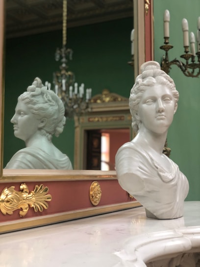 A marble bust of a woman is placed on a polished surface in front of a decorative mirror with intricate gold accents. In the background, an ornate chandelier and green wall add a classic and elegant touch to the setting.