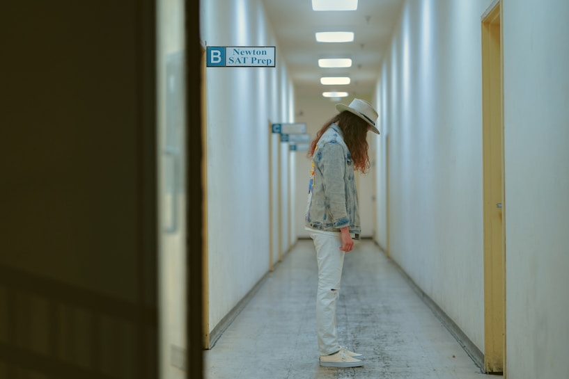 A person wearing a denim jacket, white pants, and a hat is standing in the middle of a long, narrow hallway with overhead fluorescent lights. The walls are plain and light-colored, and there are doors spaced along the corridor. A sign indicating 'Newton SAT Prep' is visible.