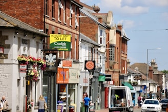 A busy street scene featuring a row of shops and businesses in buildings made of brick and stone. There are various signs for different stores and services, some with colorful awnings and flowers hanging from a wall. A van is parked on the street alongside people walking.