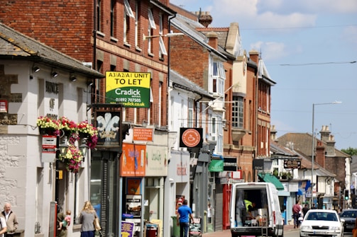 A busy street scene featuring a row of shops and businesses in buildings made of brick and stone. There are various signs for different stores and services, some with colorful awnings and flowers hanging from a wall. A van is parked on the street alongside people walking.