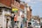 A busy street scene featuring a row of shops and businesses in buildings made of brick and stone. There are various signs for different stores and services, some with colorful awnings and flowers hanging from a wall. A van is parked on the street alongside people walking.