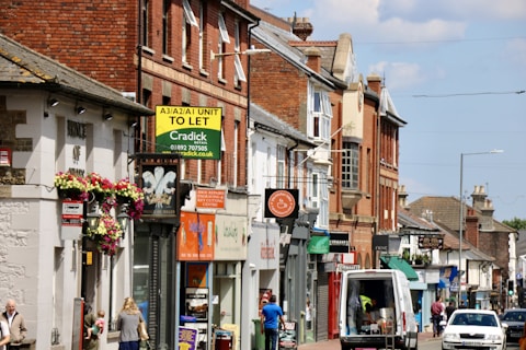 A busy street scene featuring a row of shops and businesses in buildings made of brick and stone. There are various signs for different stores and services, some with colorful awnings and flowers hanging from a wall. A van is parked on the street alongside people walking.