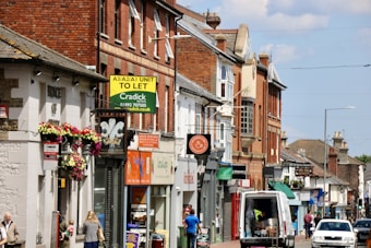 A busy street scene featuring a row of shops and businesses in buildings made of brick and stone. There are various signs for different stores and services, some with colorful awnings and flowers hanging from a wall. A van is parked on the street alongside people walking.