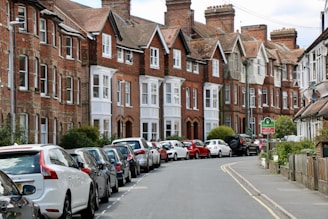 A row of traditional red-brick terraced houses line a curved street. Numerous cars are parked in an orderly fashion along the road. The scene is well-maintained, with greenery and small trees interspersed along the sidewalk. The sky is partly cloudy.