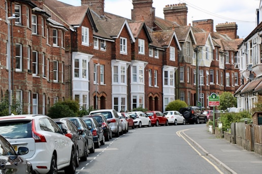 A row of traditional red-brick terraced houses line a curved street. Numerous cars are parked in an orderly fashion along the road. The scene is well-maintained, with greenery and small trees interspersed along the sidewalk. The sky is partly cloudy.