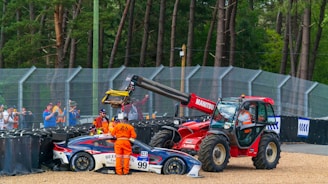 A race car with number 99 is being attended to after a crash, with several people in orange uniforms nearby. A red tractor is positioned next to the car, lifting it with its crane. Spectators watch the scene from behind a safety barrier, and the environment is surrounded by trees, indicating a racetrack in a wooded area.