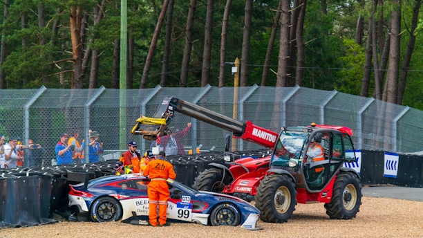 A race car with number 99 is being attended to after a crash, with several people in orange uniforms nearby. A red tractor is positioned next to the car, lifting it with its crane. Spectators watch the scene from behind a safety barrier, and the environment is surrounded by trees, indicating a racetrack in a wooded area.
