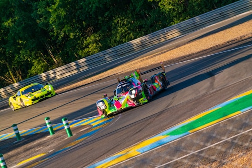 Two brightly colored racing cars speed around a track, surrounded by lush green trees and high barriers. The road is marked with yellow and blue lines, indicating a section of the circuit. The scene conveys a sense of motion and excitement.