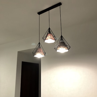 Three pendant lights with a geometric, diamond-like design hang from the ceiling. The lamps have a metallic frame enclosing the light bulbs and create an artistic lighting effect against a plain white ceiling.