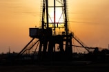 A silhouetted oil rig stands against a golden sunset. The structure's intricate details are highlighted by the backlight, creating a dramatic contrast with the glowing sky. The sun is partially visible, casting rays through the metal framework.