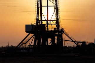A silhouetted oil rig stands against a golden sunset. The structure's intricate details are highlighted by the backlight, creating a dramatic contrast with the glowing sky. The sun is partially visible, casting rays through the metal framework.