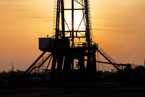 A silhouetted oil rig stands against a golden sunset. The structure's intricate details are highlighted by the backlight, creating a dramatic contrast with the glowing sky. The sun is partially visible, casting rays through the metal framework.