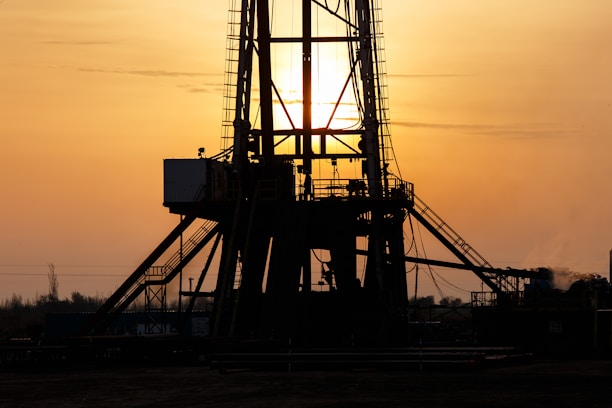 A silhouetted oil rig stands against a golden sunset. The structure's intricate details are highlighted by the backlight, creating a dramatic contrast with the glowing sky. The sun is partially visible, casting rays through the metal framework.