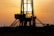 A silhouetted oil rig stands against a golden sunset. The structure's intricate details are highlighted by the backlight, creating a dramatic contrast with the glowing sky. The sun is partially visible, casting rays through the metal framework.