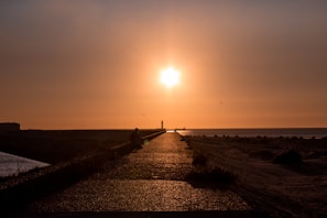 A pathway stretches towards a bright setting sun, creating a silhouette of a person sitting along the path. The sky is filled with warm orange and yellow hues, and the pathway is bordered by low vegetation and sand.