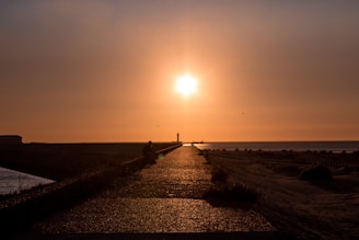 A pathway stretches towards a bright setting sun, creating a silhouette of a person sitting along the path. The sky is filled with warm orange and yellow hues, and the pathway is bordered by low vegetation and sand.