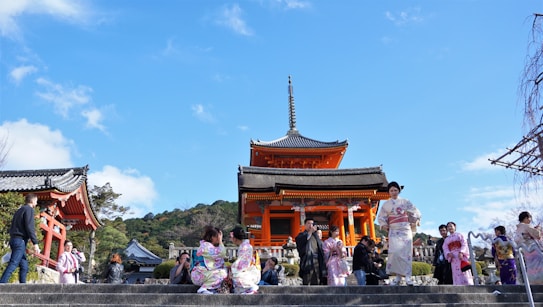 A traditional Japanese temple with vibrant orange and black accents stands under a clear blue sky. People dressed in colorful kimonos are gathered around the temple, some standing on stone steps. The scene is lively and captures a blend of historical architecture and cultural attire.