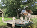 A rustic log cabin with large glass windows and a sloping roof is nestled among green trees. In front of it, a small wooden dock extends over tranquil water, where a rowboat with oars is tied. The surrounding area is lush with grass and foliage, evoking a serene, natural setting.