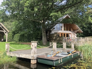 A rustic log cabin with large glass windows and a sloping roof is nestled among green trees. In front of it, a small wooden dock extends over tranquil water, where a rowboat with oars is tied. The surrounding area is lush with grass and foliage, evoking a serene, natural setting.