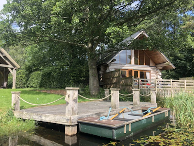A rustic log cabin with large glass windows and a sloping roof is nestled among green trees. In front of it, a small wooden dock extends over tranquil water, where a rowboat with oars is tied. The surrounding area is lush with grass and foliage, evoking a serene, natural setting.