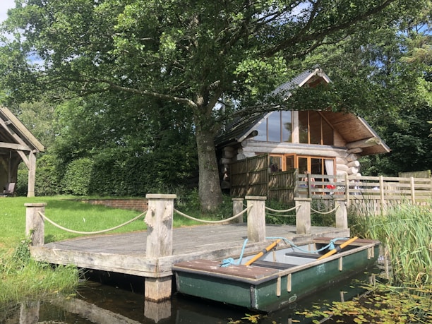A rustic log cabin with large glass windows and a sloping roof is nestled among green trees. In front of it, a small wooden dock extends over tranquil water, where a rowboat with oars is tied. The surrounding area is lush with grass and foliage, evoking a serene, natural setting.