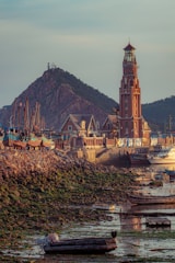 A tall lighthouse made of red bricks stands prominently near the shoreline, surrounded by several rustic buildings. Fishing boats are lined up by the coast, with hills in the background. The foreground features a small rowboat resting on a rocky and algae-covered shore, and there is soft lighting indicating early morning or late afternoon.
