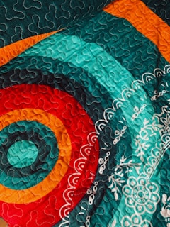 A vibrant fabric quilt with intricate patterns, featuring circular designs in red, orange, and teal. The quilt displays symmetrical patterns with swirling stitches and floral motifs in white.