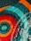 A vibrant fabric quilt with intricate patterns, featuring circular designs in red, orange, and teal. The quilt displays symmetrical patterns with swirling stitches and floral motifs in white.