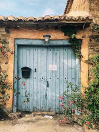 A rustic wooden door with a light blue paint finish, framed by aged stone walls and creeping vines. A black mailbox is attached to the door, and there is a small sign near its center. Pink flowers and green foliage weave around the entrance, adding a vibrant contrast to the weathered wood. A black lantern hangs above the door, providing a quaint and vintage touch.