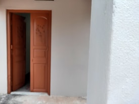 A partially open wooden door leads to an interior space, with a textured white wall on the right. The door has ornamental carvings and darker wood grain patterns.