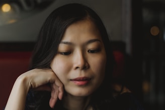 A person with long dark hair appears to be deep in thought, with their hand resting against their cheek. The background is softly blurred, with dim lighting contributing to an intimate and contemplative atmosphere.