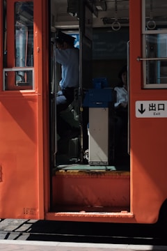 The image captures the entrance of a red public transport vehicle, likely a tram or bus. A person in a uniform, possibly the driver, is visible sitting at the front, partially obscured by shadows. Another passenger is seated further inside, also partially visible. Sunlight casts strong shadows inside the vehicle, creating a contrast between the interior and exterior.