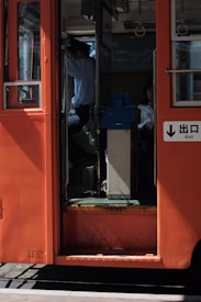 The image captures the entrance of a red public transport vehicle, likely a tram or bus. A person in a uniform, possibly the driver, is visible sitting at the front, partially obscured by shadows. Another passenger is seated further inside, also partially visible. Sunlight casts strong shadows inside the vehicle, creating a contrast between the interior and exterior.