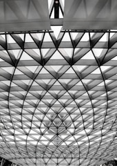 A geometric structure with intersecting triangular patterns creates a dome effect. The design consists of metal or glass panels that appear translucent, allowing a faint view of a large building or tower in the background. The structure is both modern and architecturally intriguing.