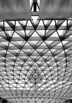 A geometric structure with intersecting triangular patterns creates a dome effect. The design consists of metal or glass panels that appear translucent, allowing a faint view of a large building or tower in the background. The structure is both modern and architecturally intriguing.