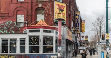 A vibrant urban street scene featuring a variety of storefronts with colorful signs in different languages. The architecture includes a prominent red brick building adorned with graffiti. A distinct structure resembling a pagoda roof is visible above the corner establishment. Pedestrians walk along the sidewalk, adding lively human activity to the scene. Numerous advertisements and signboards create an eclectic mix of visual elements typical of a bustling city neighborhood.