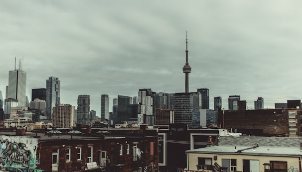 A cityscape featuring a mix of skyscrapers and mid-rise buildings under a cloudy, overcast sky. The tall, prominent CN Tower stands out in the skyline. In the foreground, older brick buildings with graffiti add an urban touch.