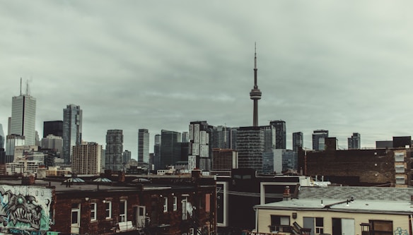 A cityscape featuring a mix of skyscrapers and mid-rise buildings under a cloudy, overcast sky. The tall, prominent CN Tower stands out in the skyline. In the foreground, older brick buildings with graffiti add an urban touch.