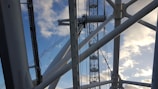 Large metal structures and cables intersect against a backdrop of a clear blue sky with scattered clouds, suggesting an architectural or engineering structure.
