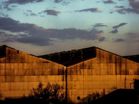 A large industrial warehouse building is illuminated by warm, golden light, casting shadows that accentuate its corrugated metal texture. The sky above displays a mix of blue and soft grey clouds, creating a contrast with the rigid structure below. In the foreground, silhouettes of sparse trees and utility poles add depth to the scene.