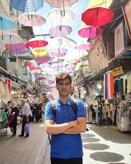 A young man, with a backpack, stands confidently with crossed arms in a busy market street. Vibrantly colored umbrellas are suspended overhead, creating a lively and shaded atmosphere. The market is bustling with people walking and shopping. Various shops lining the street display colorful clothing and textiles.