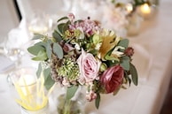 A bouquet of flowers featuring a variety of blooms, including roses, surrounded by eucalyptus leaves. The arrangement is placed on a white table, with softly blurred background elements such as glasses and candles giving a warm and elegant ambiance.