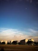 A serene landscape featuring several houses positioned in the foreground against a vast, open field. The sky is a rich gradient of blue transitioning to lighter shades near the horizon, with a small moon visible in the upper part.