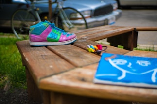 A colorful sneaker with pink, green, and blue sections is placed on a wooden bench. In the foreground, there are small paint bottles and a vibrant blue artwork depicting a simplistic human face. The background includes a bicycle and a parked car, slightly out of focus, suggesting an outdoor setting.