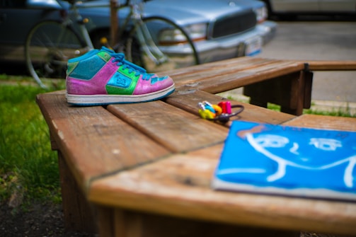 A colorful sneaker with pink, green, and blue sections is placed on a wooden bench. In the foreground, there are small paint bottles and a vibrant blue artwork depicting a simplistic human face. The background includes a bicycle and a parked car, slightly out of focus, suggesting an outdoor setting.