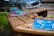 A colorful sneaker with pink, green, and blue sections is placed on a wooden bench. In the foreground, there are small paint bottles and a vibrant blue artwork depicting a simplistic human face. The background includes a bicycle and a parked car, slightly out of focus, suggesting an outdoor setting.